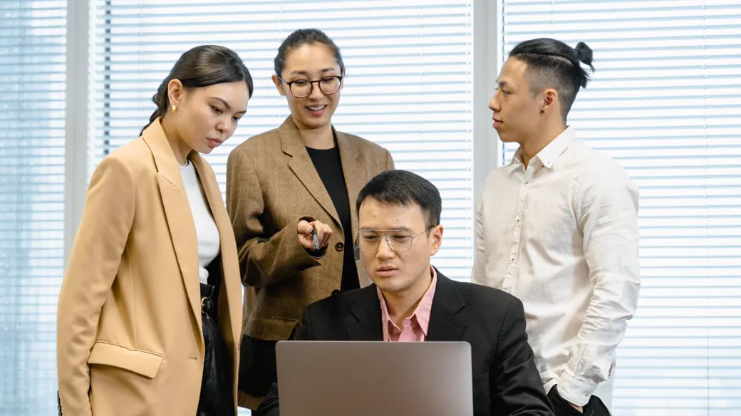 Nhóm nhân viên văn phòng quây quanh đồng nghiệp dùng laptop, hình ảnh công ty nhiều người cùng vận hành