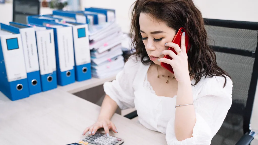 Nữ chủ cửa hàng nghe điện thoại vừa bấm máy tính, phía sau là kệ bìa hồ sơ, tính doanh thu để xác định thuế