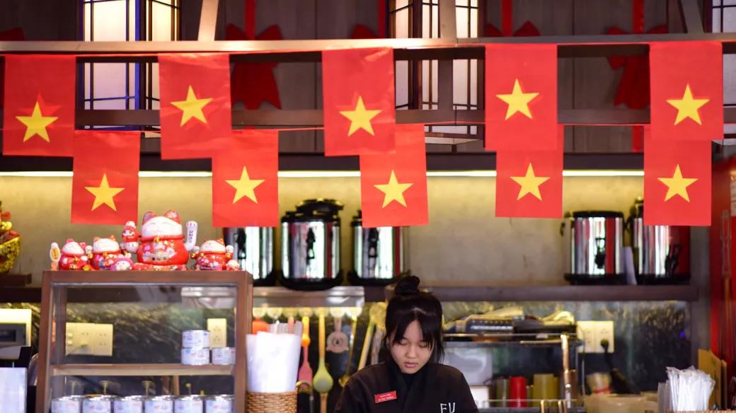 Quầy bán đồ ăn uống treo cờ đỏ sao vàng, nhân viên đứng sau quầy, hộ kinh doanh có nhiều giao dịch nhỏ